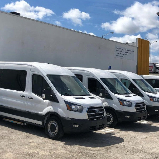 A row of white Ford Transit vans parked in an outdoor lot under a blue sky with clouds, with a large white building in the background.